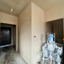 Wnętrze w trakcie prac wykończeniowych: świeżo nałożona warstwa zaprawy na ścianach, widoczny agregat tynkarski z niebieskim silnikiem oraz fragmenty instalacji elektrycznej wystające ze ściany...