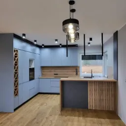 Jasnoniebieska kuchnia z drewnianymi akcentami, wyspą z czarnym panelem i oryginalnym oświetleniem sufitowym