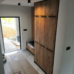 Wysoka szafa z drewnopodobnymi frontami i czarnymi detalami, wkomponowana we wnękę przy wejściu, z siedziskiem obok, na podłodze karton zabezpieczający.