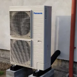 Zewnętrzna jednostka pompy ciepła Panasonic, zamontowana na betonowych podstawach przy ścianie budynku, z widoczną rurą odprowadzającą skropliny i czerwoną rurą spustową.