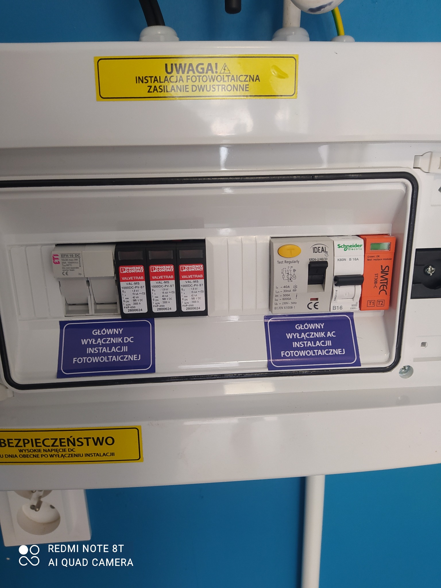 Rozdzielnica elektryczna z wyłącznikami instalacji fotowoltaicznej DC i AC, z żółtymi naklejkami ostrzegawczymi o napięciu i zasilaniu dwustronnym.