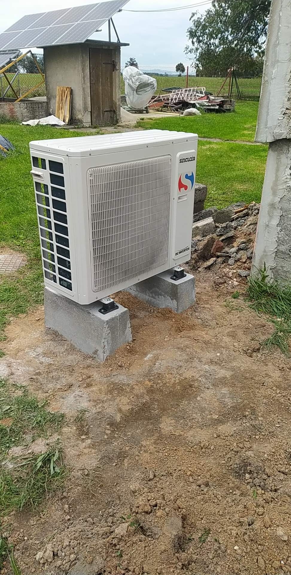 Biała jednostka zewnętrzna pompy ciepła marki Sinclair, zamontowana na betonowych bloczkach, z widocznym logo firmy i panele słoneczne w tle na trawiastym terenie.