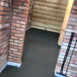 Świeżo wylana, gładka, szara posadzka w pomieszczeniu o ścianach z czerwonej cegły i drewnianych bali.