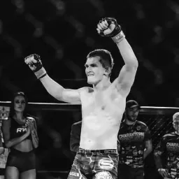 Czarno-białe zdjęcie młodego zawodnika sportów walki z uniesionymi rękami w geście zwycięstwa, w tle publiczność i ring.