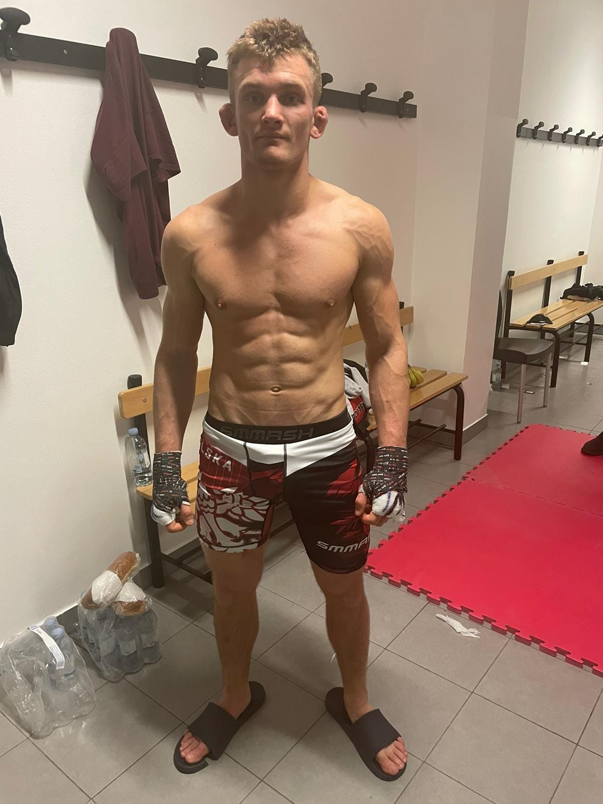 Młody, wysportowany mężczyzna z zaklejonymi dłońmi, w spodenkach do sportów walki, stojący na sali treningowej z matą na podłodze. Widoczne zniekształcone uszy, prawdopodobnie po treningach. W tle...