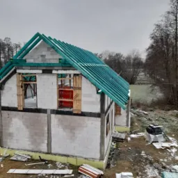 Wznoszony, dwukondygnacyjny budynek z bloczków, z zieloną konstrukcją dachu bez pokrycia, zabezpieczonymi oknami, w zimowej scenerii.
