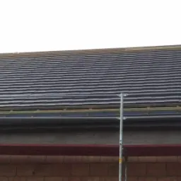 Fragment dachu z ciemną dachówką w trakcie układania, widoczne rusztowanie i elementy konstrukcji drewnianej pod dachówką.