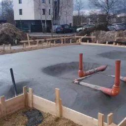 Zalana betonem płyta fundamentowa z wystającymi pionowo rurami kanalizacyjnymi, otoczona drewnianym szalunkiem, na tle placu budowy i bloku mieszkalnego.