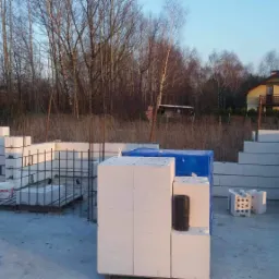 Wznoszenie ścian z białych bloczków silikatowych na fundamencie zbrojonym stalą; widoczne elementy izolacji i paleta z bloczkami pod plandeką, w tle dom jednorodzinny i drzewa.