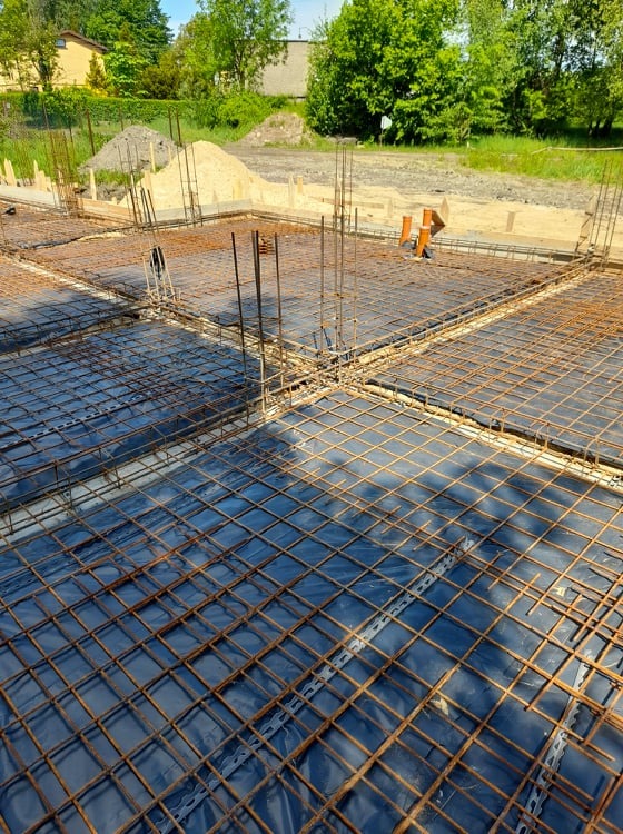 Zbrojenie płyty fundamentowej z wystającymi prętami pionowymi, widok z góry na plac budowy z hałdą ziemi w tle.