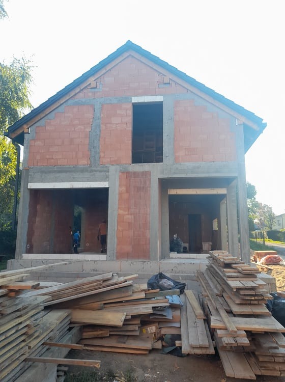 Dwukondygnacyjny budynek w stanie surowym otwartym, z widocznymi czerwonymi bloczkami i betonowymi słupami nośnymi, z deskami leżącymi na pierwszym planie.