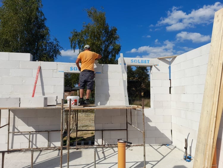 Pracownik w pomarańczowej koszulce muruje ściany z białych bloczków Solbet na budowie domu, widoczne rusztowanie, poziomica i niebo z chmurami.