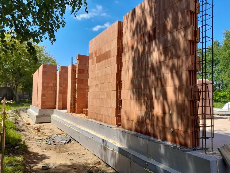 Wznoszenie ścian z czerwonej cegły na fundamencie ocieplonym styropianem, widoczne zbrojenie pionowe, w tle drzewa.