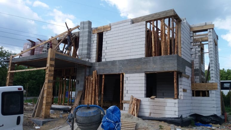 Budowa dwukondygnacyjnego domu z białych bloczków, widoczne drewniane szalunki i podpory, pracownik na rusztowaniu, betoniarka i materiały budowlane w tle.