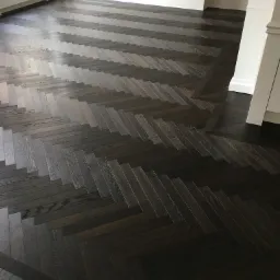 Ciemna, lakierowana podłoga ułożona w jodełkę francuską, z widocznym fragmentem białej listwy przypodłogowej i grzejnika pod oknem.