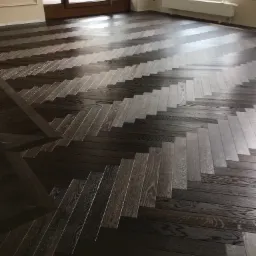 Ciemna, drewniana podłoga ułożona w jodełkę, z widocznymi słojami drewna i refleksami światła, w tle okno balkonowe i grzejnik.