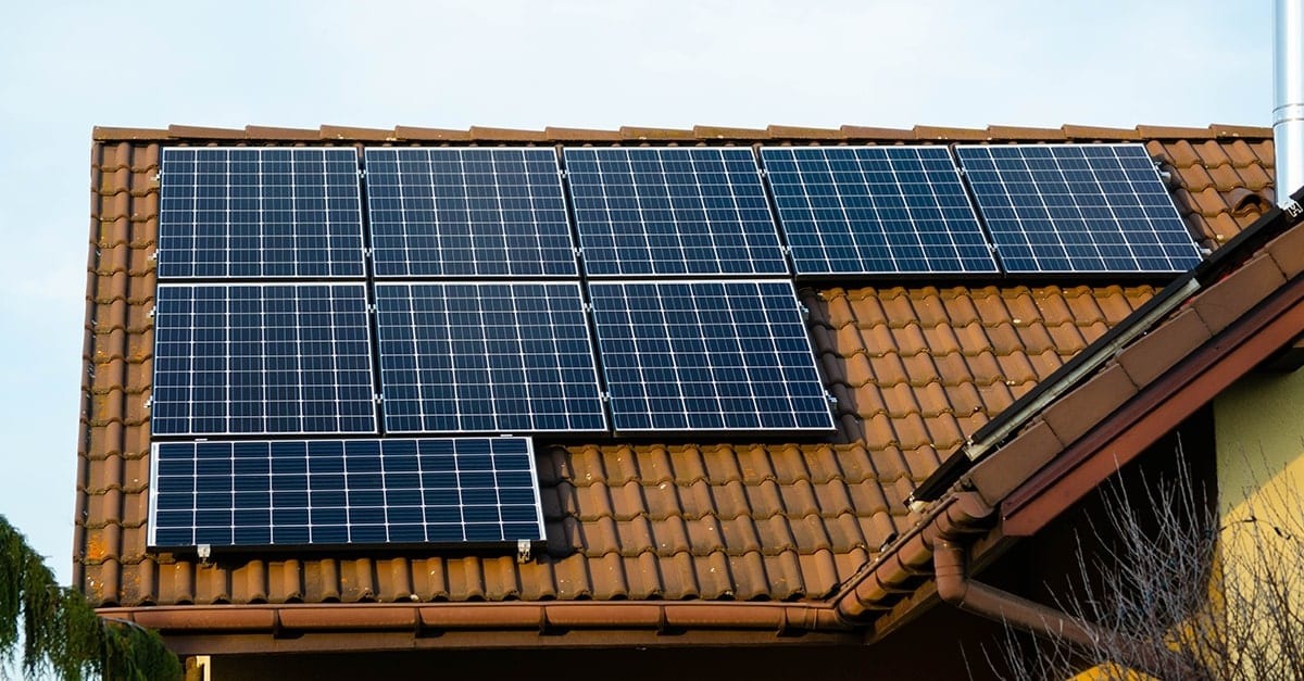 Panele słoneczne zamontowane na dachu z brązowej dachówki, widoczne rury spustowe i komin ze stali nierdzewnej.