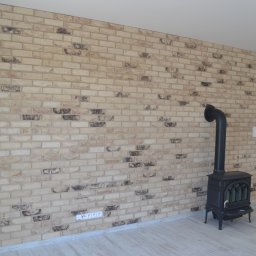 RIEMCHENBAU sp. z o.o. - Ściana wykończona cegłą klinkierową w odcieniach beżu i brązu z widocznym kominkiem wolnostojącym w salonie z jasną podłogą.