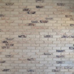 RIEMCHENBAU sp. z o.o. - Ściana wewnętrzna wykończona dekoracyjną cegłą w odcieniach beżu i brązu, z widocznymi fugami, oświetlona naturalnym światłem, z fragmentem drewnianej ościeżnicy po lewej stronie i białym oknem po...