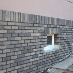 RIEMCHENBAU sp. z o.o. - Nowa elewacja z szarej cegły klinkierowej wokół okien piwnicznych, widoczne detale wykończenia i izolacja przy gruncie.