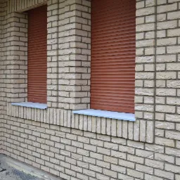 Elewacja z jasnej cegły klinkierowej z dwoma oknami zabezpieczonymi brązowymi roletami zewnętrznymi, widoczne detale wykończenia wokół okien i parapety.