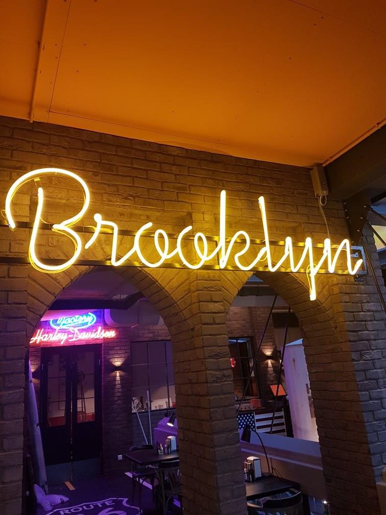 Neonowy napis 'Brooklyn' nad ceglanym łukiem wejścia do lokalu, w tle widoczny neon Harley-Davidson.