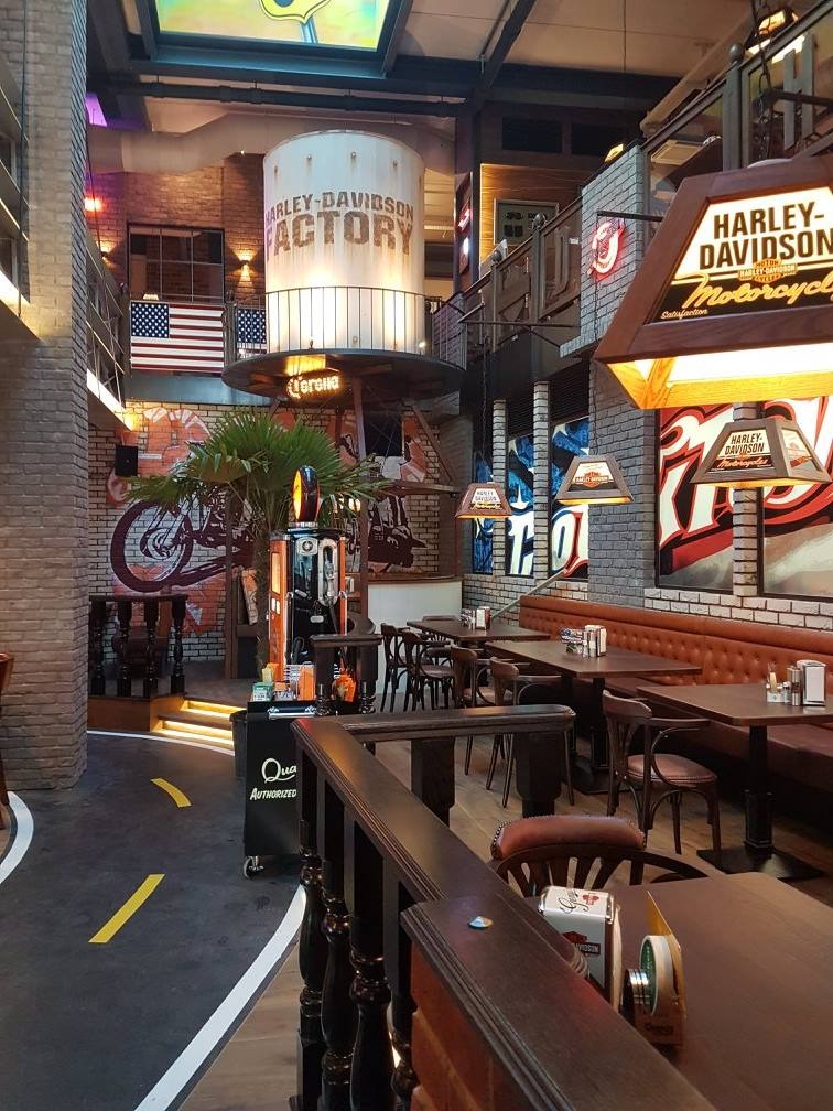 Wnętrze restauracji w stylu industrialnym z motywami Harley-Davidson, widoczne ceglane ściany, malowidła motocykli, amerykańskie flagi, oświetlenie tematyczne i meble z ciemnego drewna.