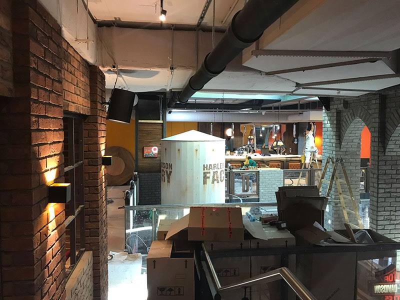 Wnętrze w trakcie wykańczania z elementami cegły na ścianach, widoczny zbiornik z napisem Harley Factory, kartony i drabina w tle.