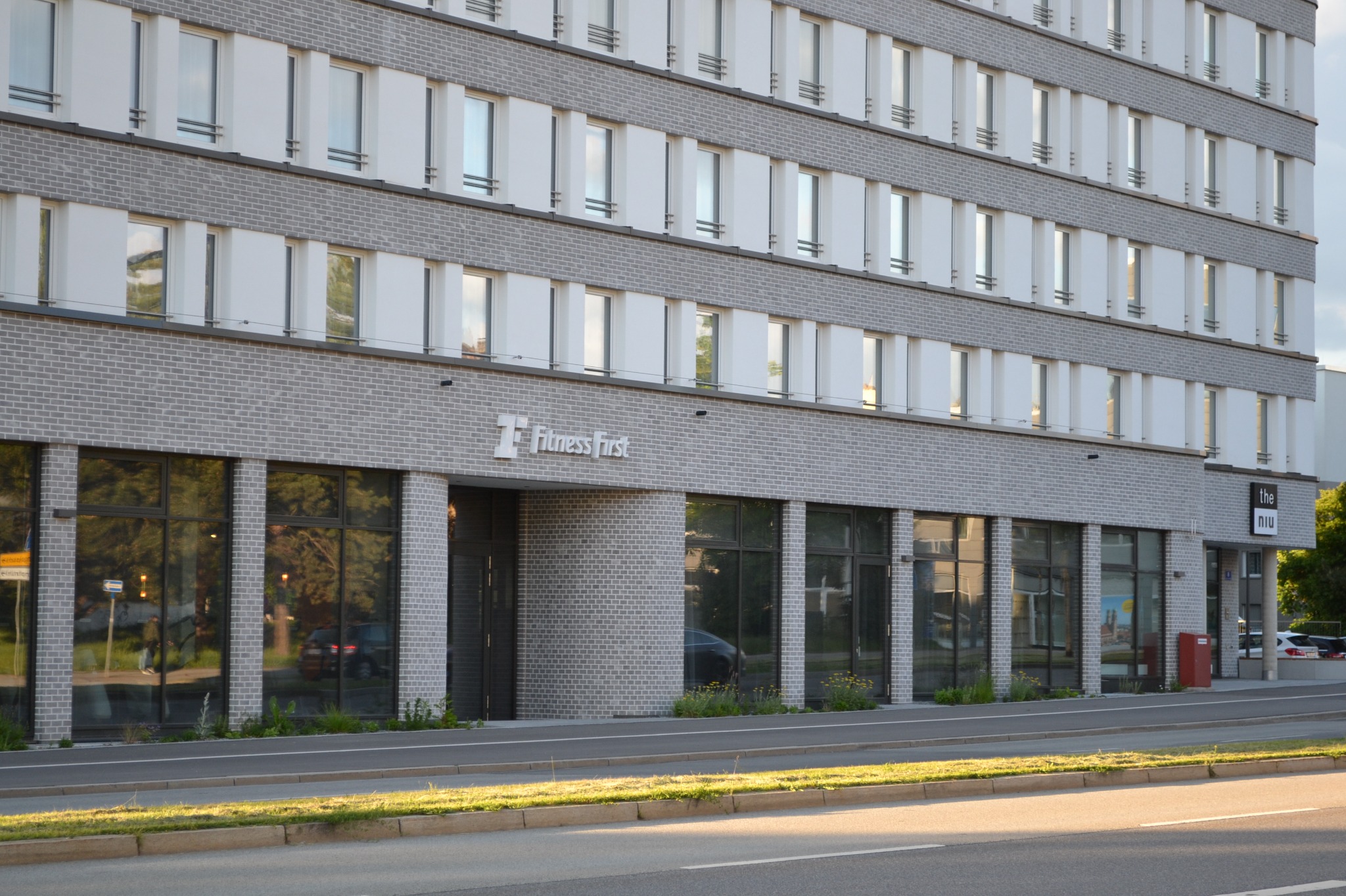 Nowoczesny budynek biurowy z szarej cegły z dużymi oknami, logo 'Fitness First' i 'the niu'