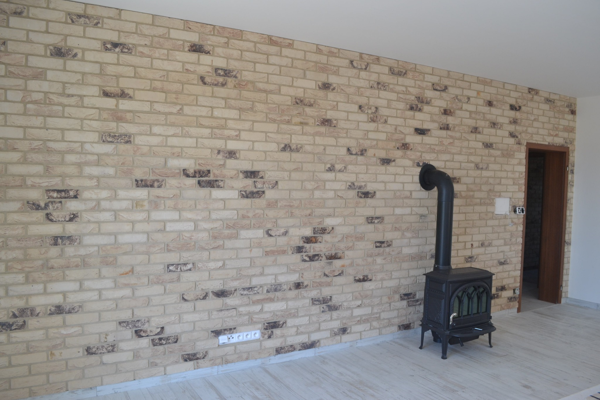 Ściana wykończona cegłą klinkierową w odcieniach beżu i brązu z widocznym kominkiem wolnostojącym w salonie z jasną podłogą.
