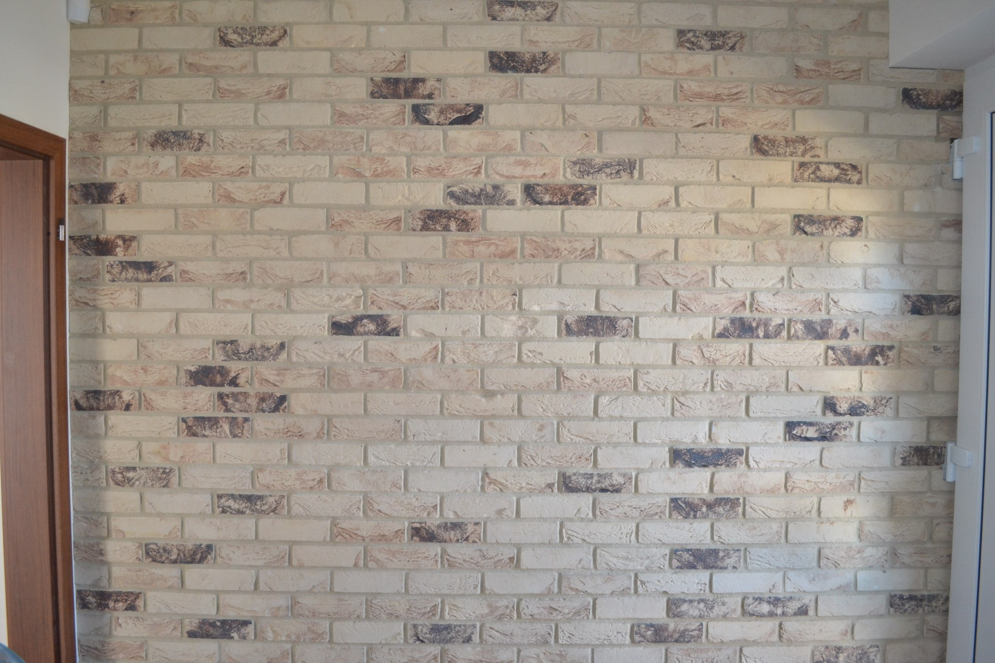 Ściana wewnętrzna wykończona dekoracyjną cegłą w odcieniach beżu i brązu, z widocznymi fugami, oświetlona naturalnym światłem, z fragmentem drewnianej ościeżnicy po lewej stronie i białym oknem po...