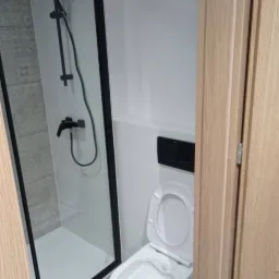 Nowoczesna łazienka z kabiną prysznicową typu walk-in z czarnymi profilami i białą ceramiką, widoczna częściowo drewniana ościeżnica drzwi