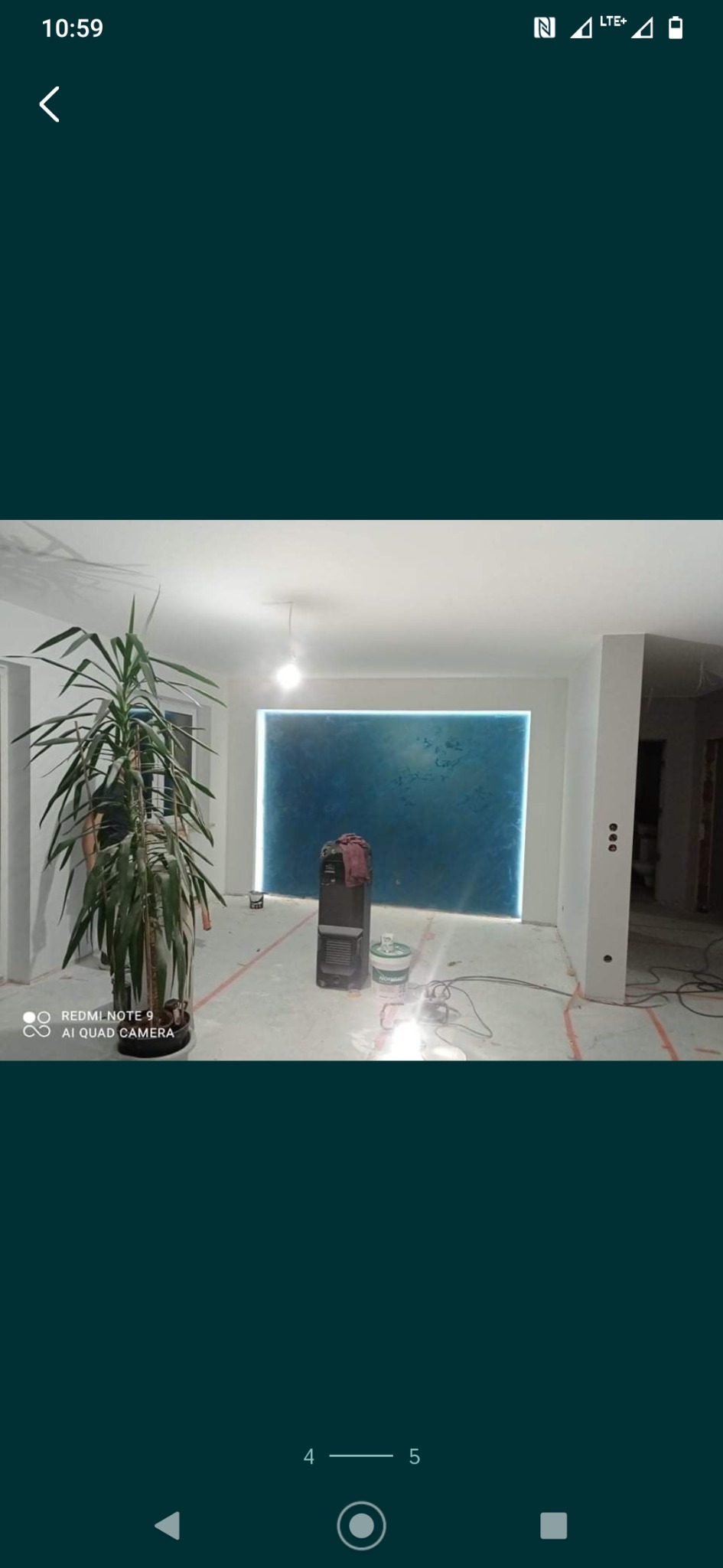 Pokój w trakcie malowania z dekoracyjną, podświetloną na niebiesko ścianą, widoczna roślina doniczkowa, grzejnik elektryczny, wiadro z farbą i narzędzia malarskie na podłodze zabezpieczonej folią.