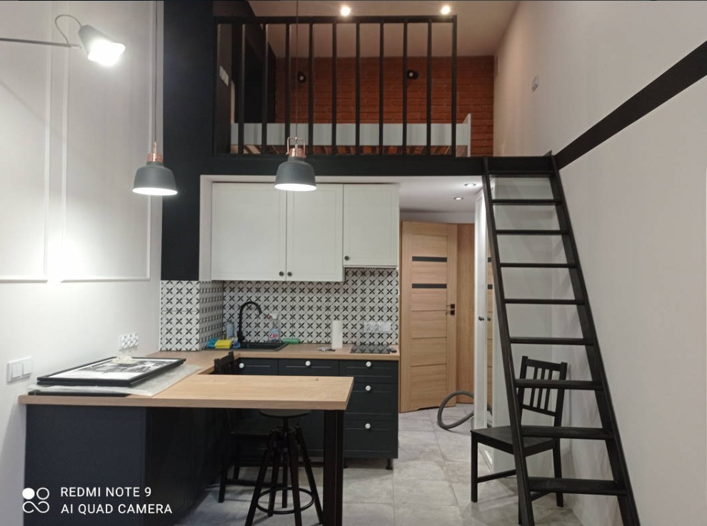 Wnętrze mieszkania z aneksem kuchennym, antresolą z barierką i drabinką, biało-czarna kolorystyka, wzorzysta mozaika nad blatem, wiszące lampy, drewniany blat baru.