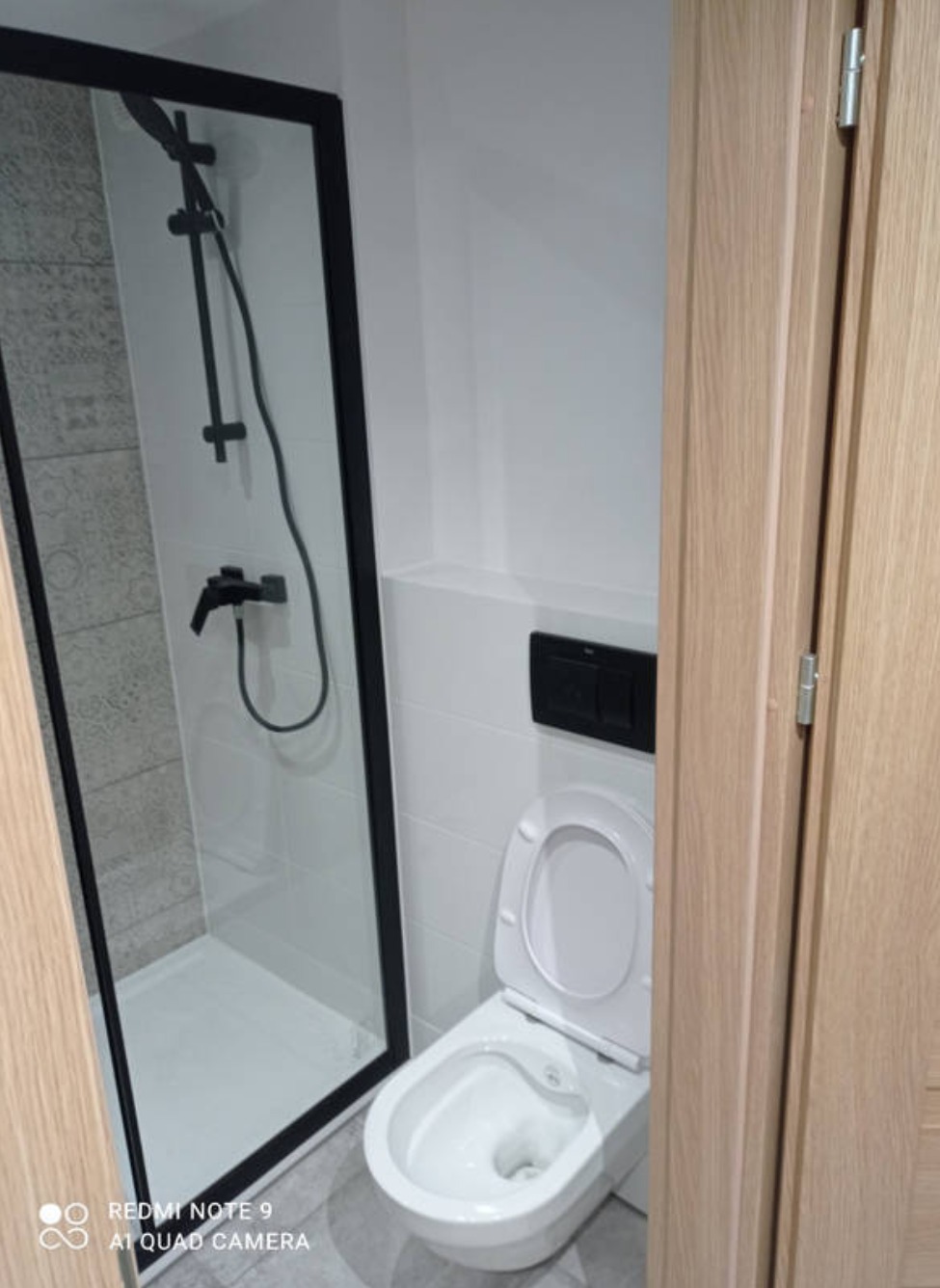 Nowoczesna łazienka z kabiną prysznicową typu walk-in z czarnymi profilami i białą ceramiką, widoczna częściowo drewniana ościeżnica drzwi