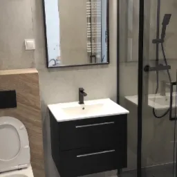 Nowoczesna łazienka z szarymi płytkami, czarną szafką pod umywalką z białą umywalką, toaletą i kabiną prysznicową z czarnymi ramami, odbicie grzejnika drabinkowego w lustrze.