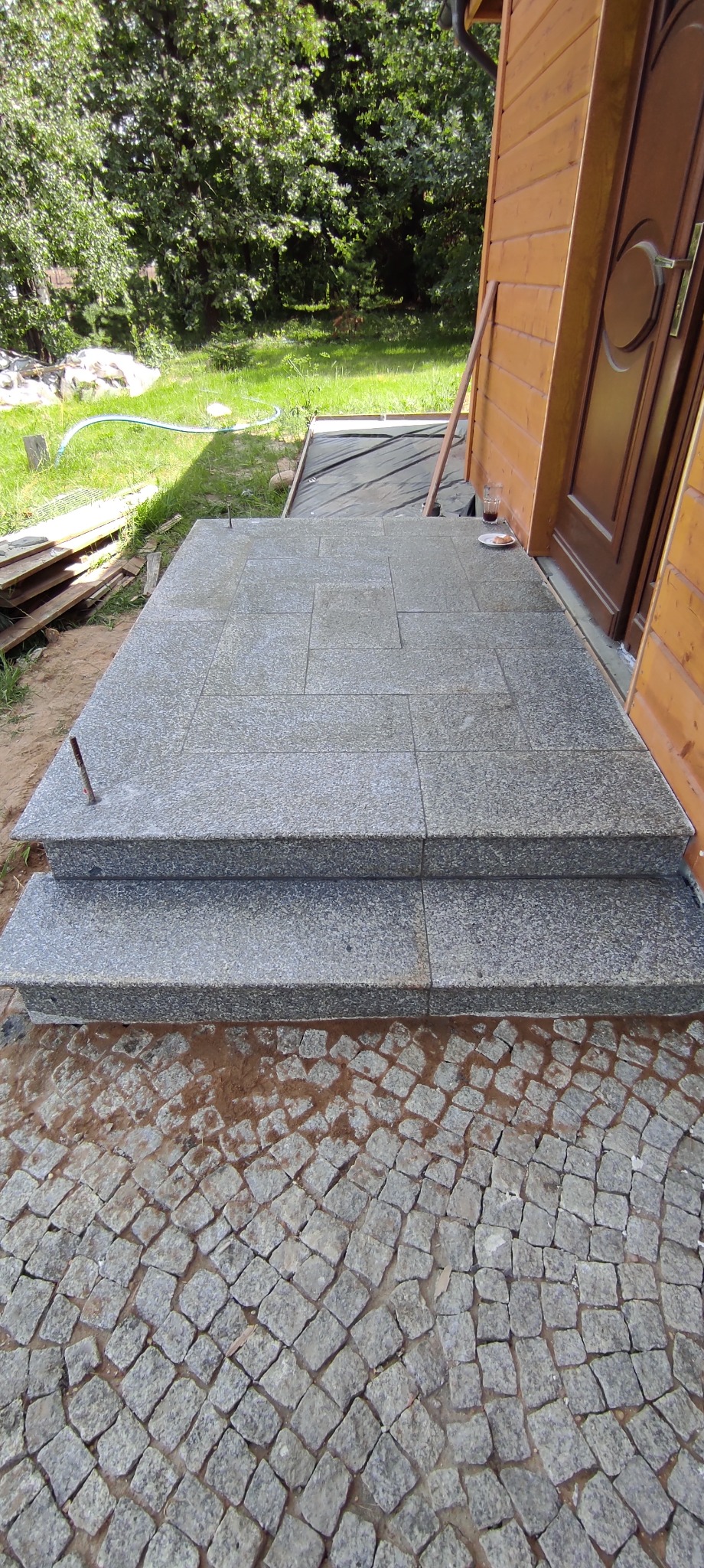 Wykończone granitowe schody wejściowe do drewnianego domu w Białymstoku, z kostką brukową na podłożu i widoczną szklanką oraz talerzykiem przy drzwiach.