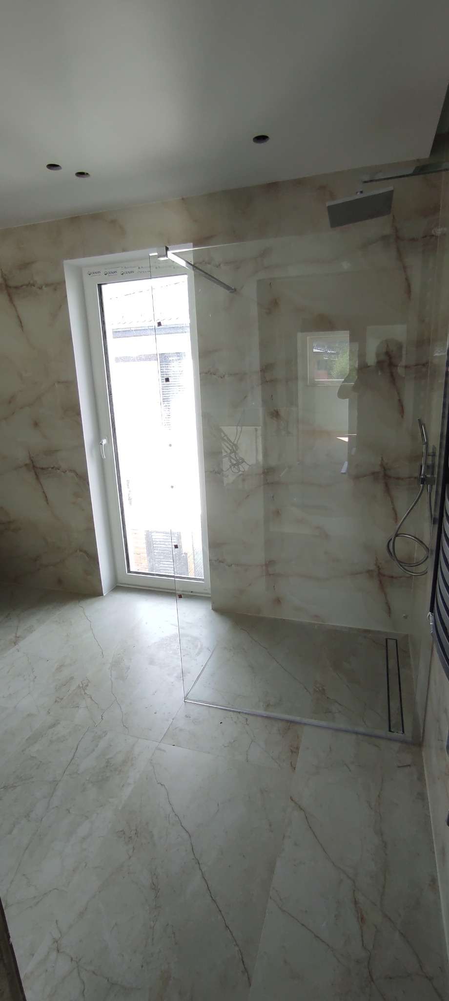 Wykończona łazienka z kabiną prysznicową bez brodzika, marmurowe płytki na ścianach i podłodze, okno z białymi ramami w pobliżu kabiny.