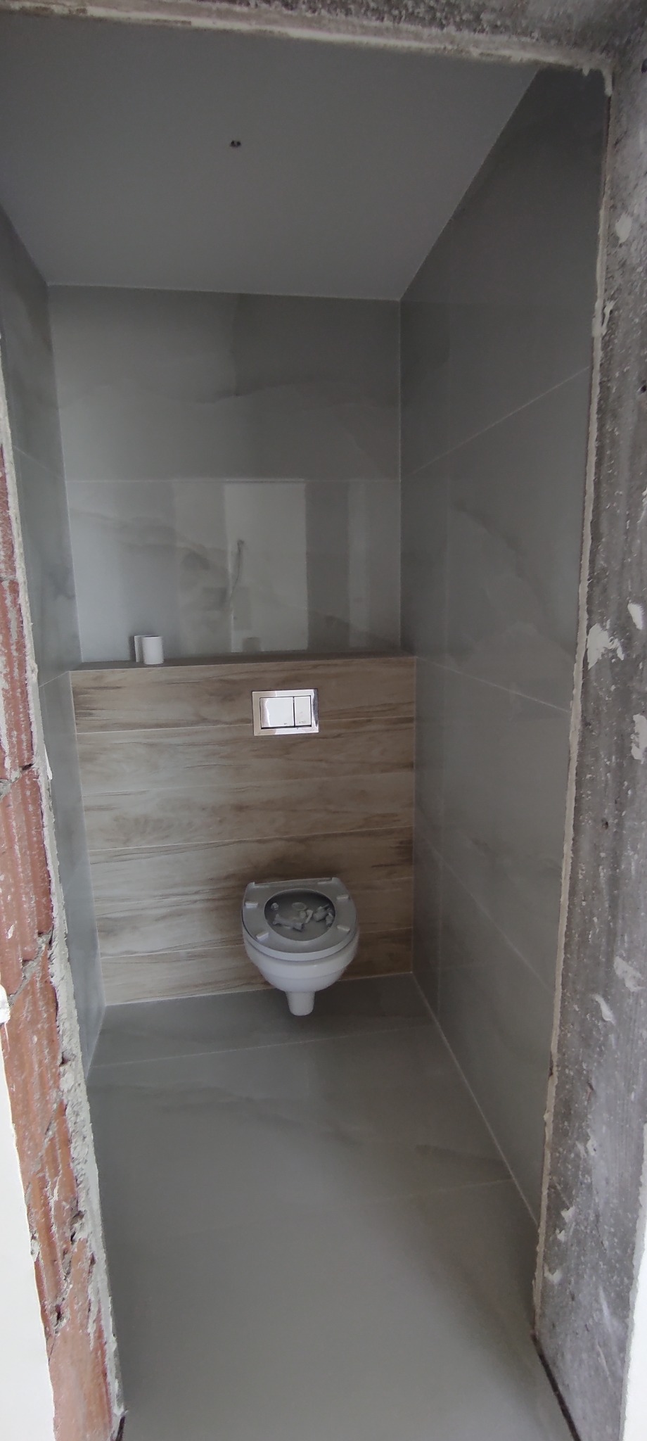 Wnętrze łazienki w trakcie prac wykończeniowych; widoczne szare płytki imitujące kamień na ścianach i podłodze, podwieszana toaleta z chromowanym przyciskiem spłukiwania oraz surowy, nieotynkowany...