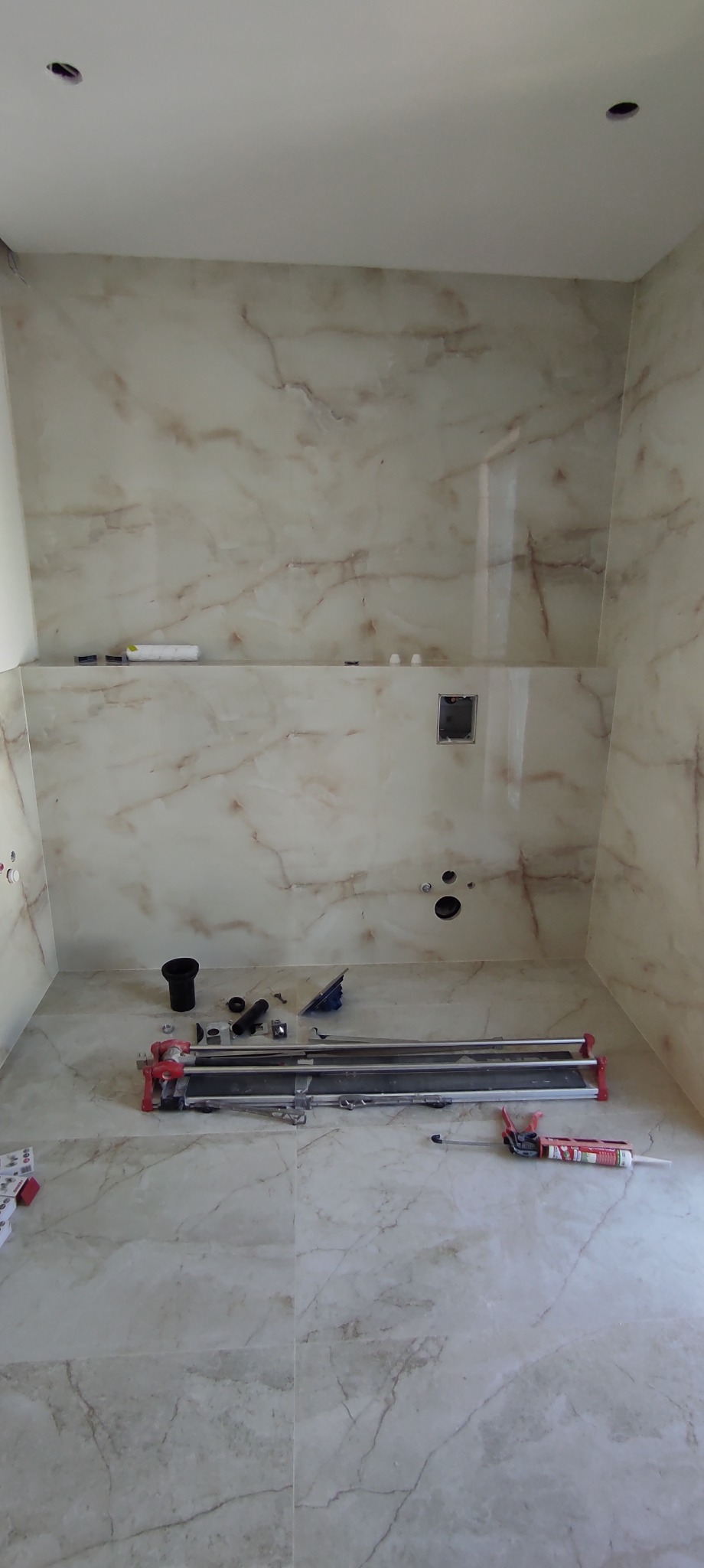 Wnętrze łazienki w trakcie prac wykończeniowych, widoczne płytki imitujące marmur na ścianach i podłodze, narzędzia glazurnicze, puszka instalacyjna i przygotowane otwory na armaturę.