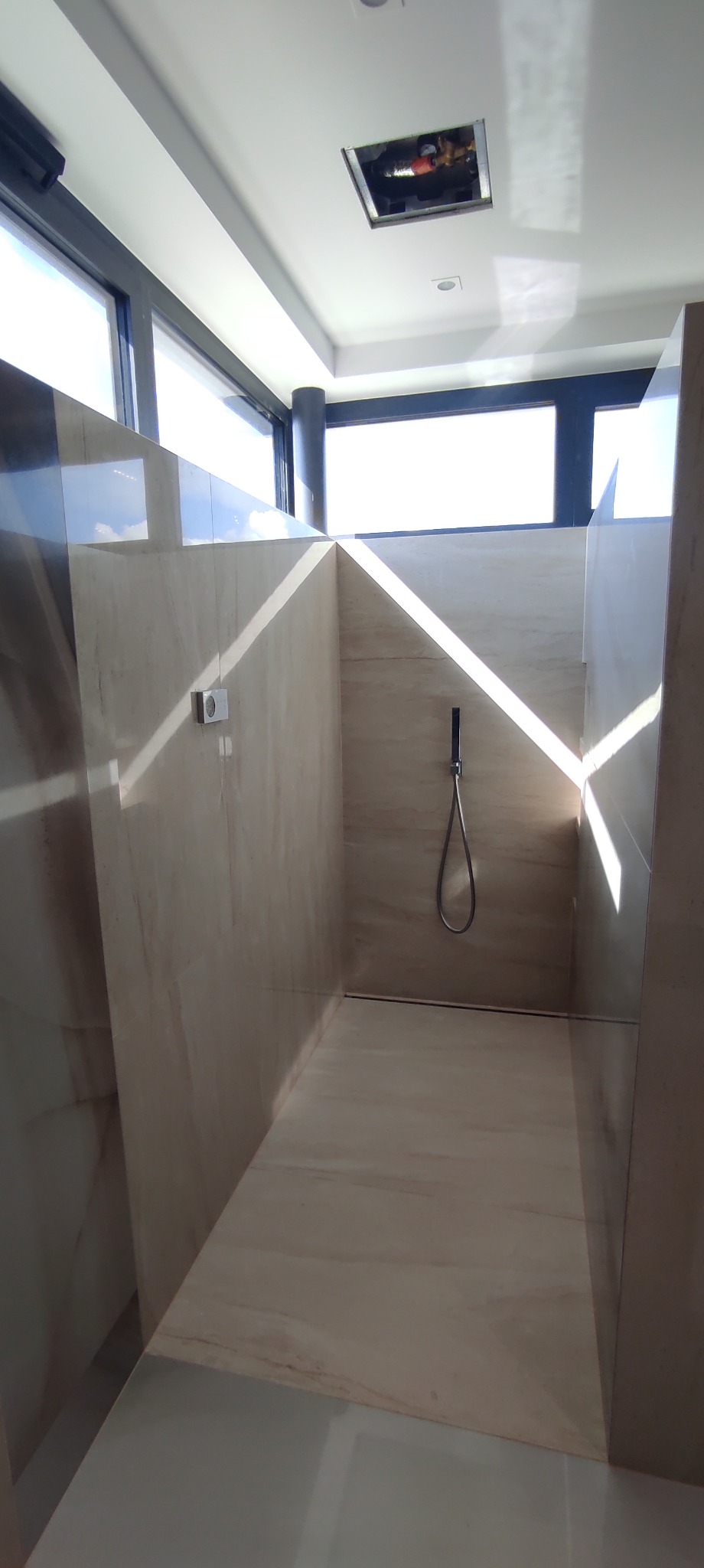 Nowoczesna kabina prysznicowa wyłożona beżowymi płytkami imitującymi marmur, z zamontowaną deszczownicą i odpływem liniowym, oświetlona naturalnym światłem wpadającym przez duże okno.