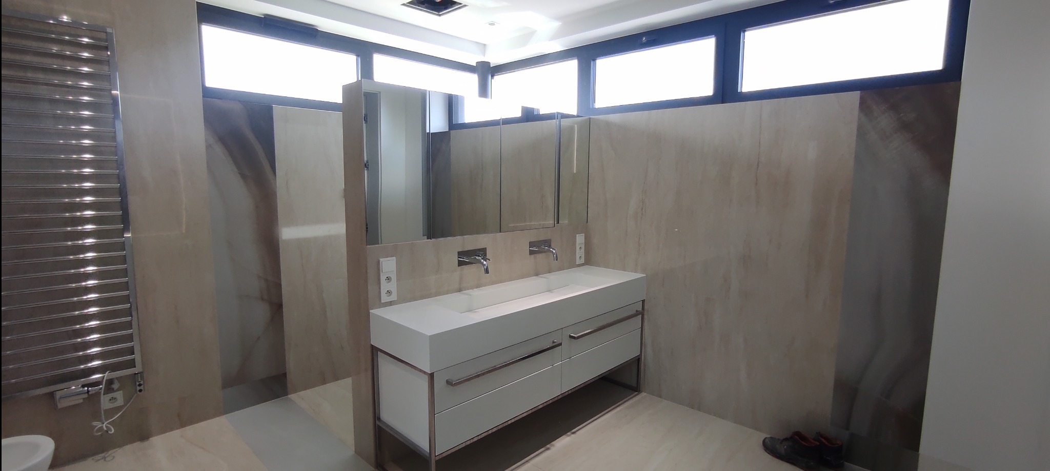 Nowoczesna łazienka z podwójną umywalką na szafce z szufladami, grzejnikiem drabinkowym i marmurowymi płytkami na ścianach, oświetlona światłem dziennym z okien pod sufitem.