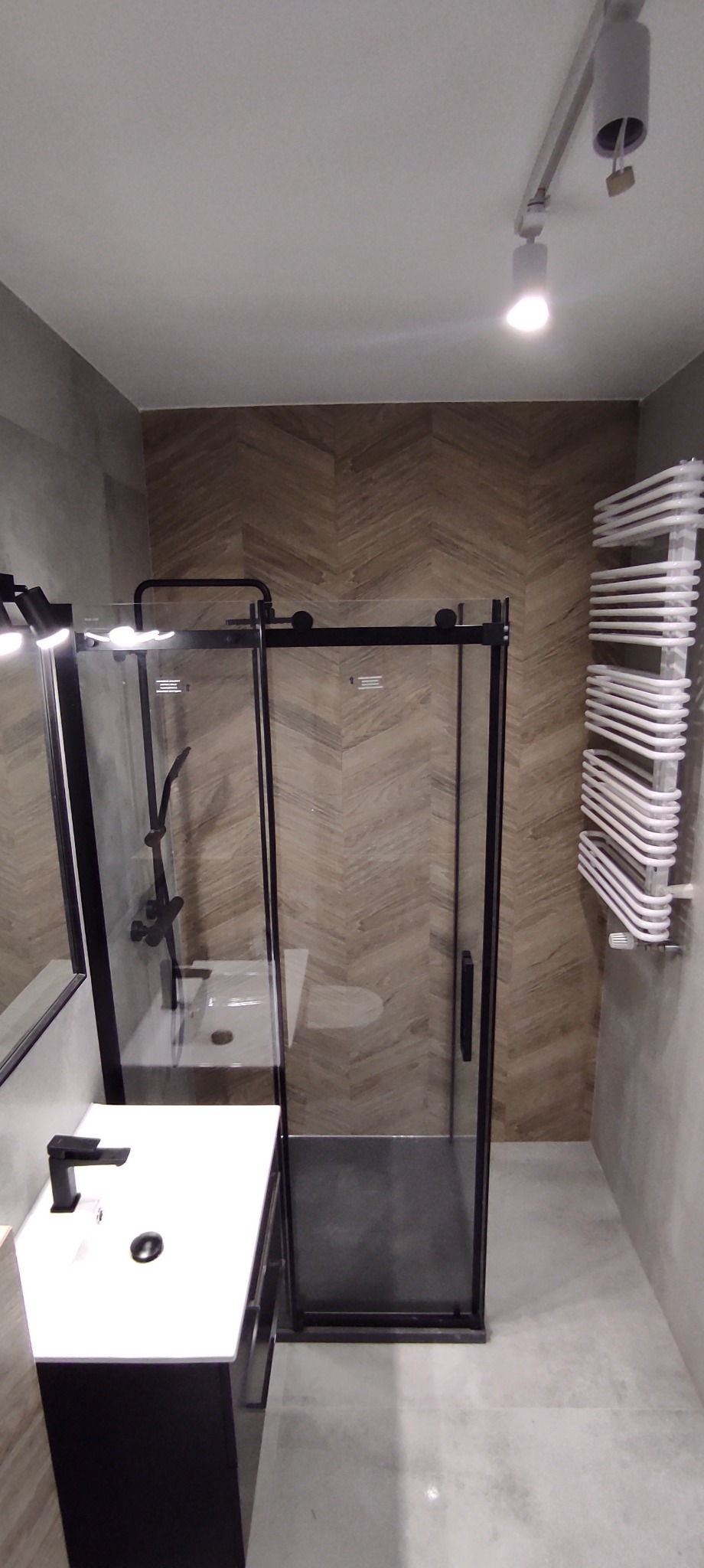 Nowoczesna łazienka z kabiną prysznicową typu walk-in z czarnymi profilami, umywalką nablatową z czarną armaturą i grzejnikiem drabinkowym, wykończona płytkami w jodełkę.