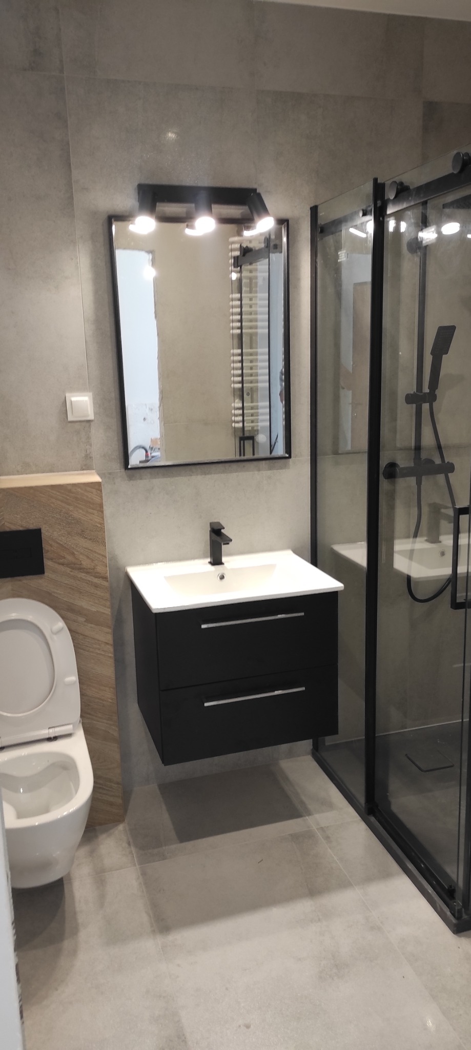 Nowoczesna łazienka z szarymi płytkami, czarną szafką pod umywalką z białą umywalką, toaletą i kabiną prysznicową z czarnymi ramami, odbicie grzejnika drabinkowego w lustrze.