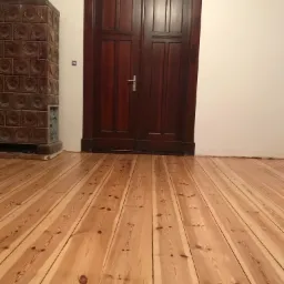 Jasne drewniane deski podłogowe po cyklinowaniu, widoczny piec kaflowy i ciemne drewniane drzwi.