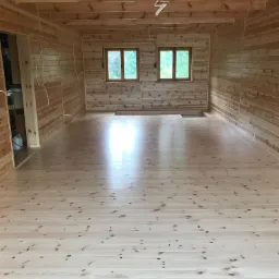 Jasne, drewniane wnętrze z wykończonymi ścianami i podłogą z desek. Dwa okna z widokiem na zieleń. Widoczne kable elektryczne zwisające z sufitu.