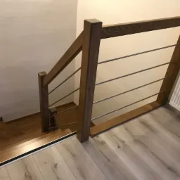 Widok z góry na drewniane schody z metalowymi poręczami, łączące podłogę wyłożoną panelami w odcieniach szarości. Grzejnik widoczny w rogu.