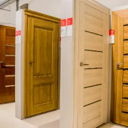 Ekspozycja różnorodnych modeli drzwi w salonie, widoczne drzwi w odcieniach drewna z różnymi wzorami i wykończeniami, niektóre z etykietami cenowymi i informacyjnymi.