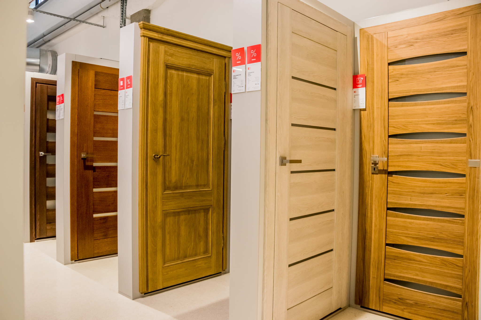 Ekspozycja różnorodnych modeli drzwi w salonie, widoczne drzwi w odcieniach drewna z różnymi wzorami i wykończeniami, niektóre z etykietami cenowymi i informacyjnymi.