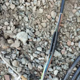 Odsłonięte przewody elektryczne z kolorowymi izolacjami, częściowo zabezpieczone czarną taśmą izolacyjną, leżą na żwirowym podłożu, obok widoczny element instalacji elektrycznej z brązową nakładką.